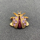 A gold ladybug-shaped brooch or charm with purple rhinestones and a dark purple center, shown on a textured gray background.