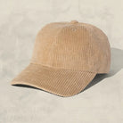 A light tan corduroy baseball cap with a curved brim, photographed on a textured light beige background with a soft shadow.