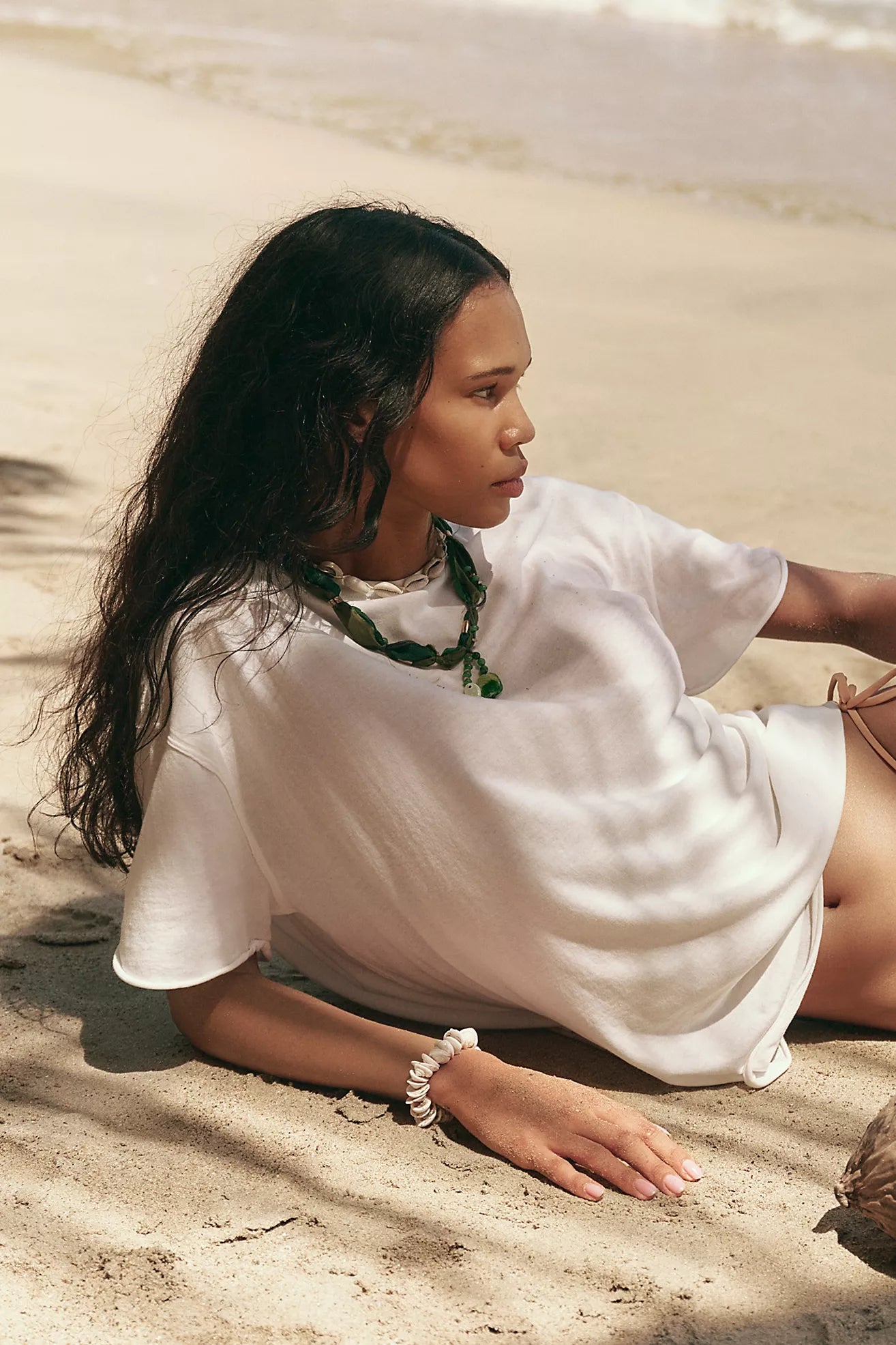 Woman sitting on sand wearing a white outfit with a green necklace