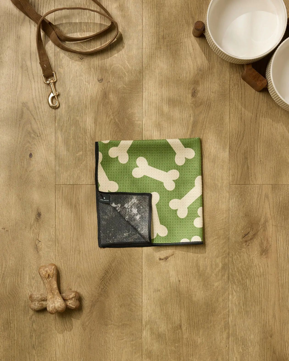 Park Run paw towel in green with cream dog bone print folded on a wooden surface with a leash and dog bowls