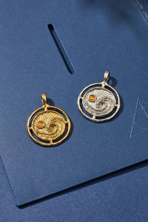 Two circular pendants, one gold and one silver with a red gemstone, on a blue background.