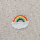 An embroidered patch featuring a small rainbow arching over a fluffy white cloud, displayed on a textured beige linen background.