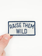 Patch with 'Raise Them Wild' text held by a hand on a white background