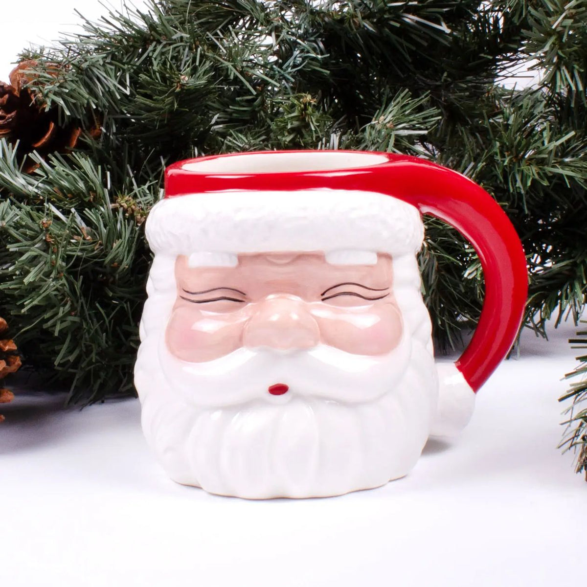 Red ceramic Santa face coffee mug with a red Santa hat, white beard, and red handle in front of evergreen branches
