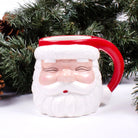 Red ceramic Santa face coffee mug with a red Santa hat, white beard, and red handle in front of evergreen branches