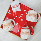 Red and white ceramic Santa mugs on a red tray with mini marshmallows, candy canes, and hot cocoa