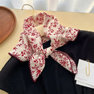 A white scarf with a red floral and botanical print, tied in a bow and draped over a black garment on a beige tabletop. A gold hanger, a wooden tray, and a paper tag are visible in the scene.