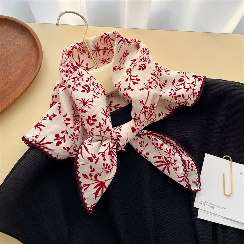 A white scarf with a red floral and botanical print, tied in a bow and draped over a black garment on a beige tabletop. A gold hanger, a wooden tray, and a paper tag are visible in the scene.