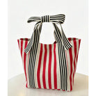 A woven tote bag with vertical red, off-white, and black stripes, featuring two top handles tied into a bow, set against a clean white background.