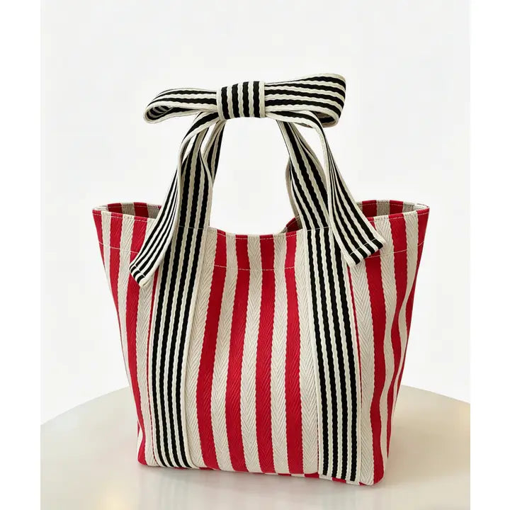 A woven tote bag with vertical red, off-white, and black stripes, featuring two top handles tied into a bow, set against a clean white background.