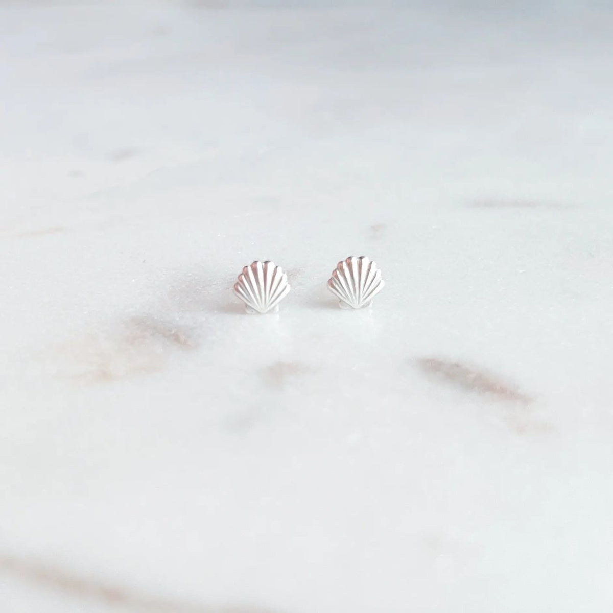 A pair of small silver stud earrings shaped like scallop shells, resting on a light marble surface.