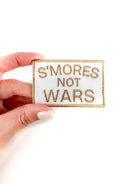 Patch with 'S'mores Not Wars' text held by a hand on a white background