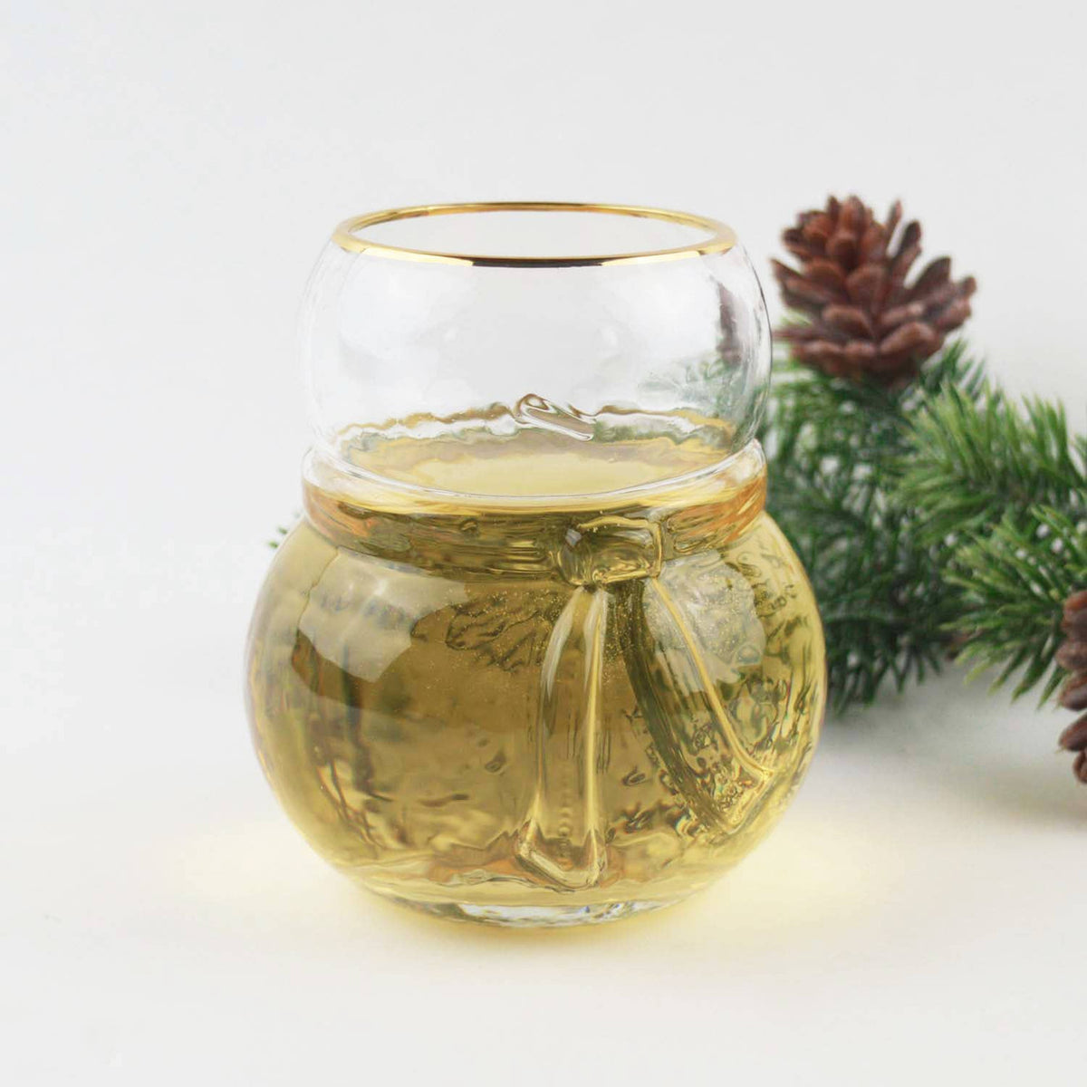 Snowman shaped gold-rimmed drinking glass filled with white wine beside evergreen and a pinecone