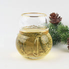 Snowman shaped gold-rimmed drinking glass filled with white wine beside evergreen and a pinecone