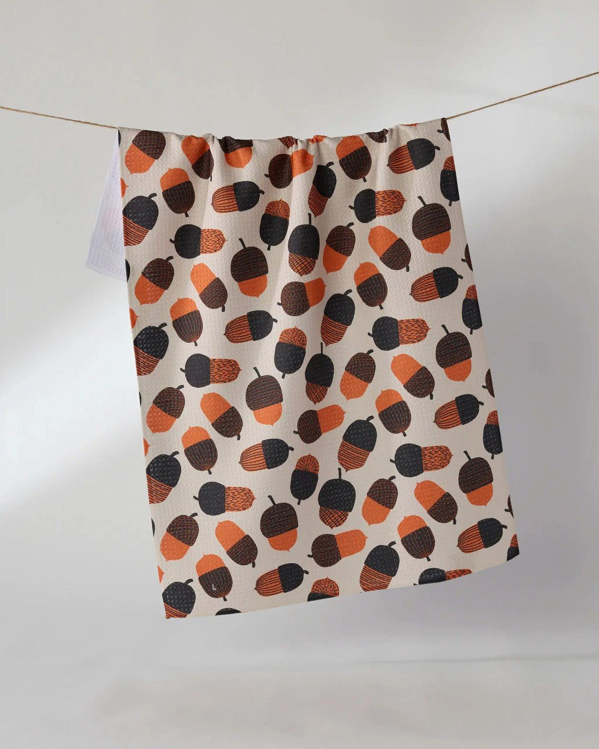 Squirrel Bounty tea towel in soft cream with orange, brown, and charcoal acorn print hanging on a line