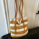 A woven straw tote bag with horizontal mustard yellow and cream stripes, featuring tan faux-leather shoulder straps, resting on a black leather sofa in front of a white paneled door.