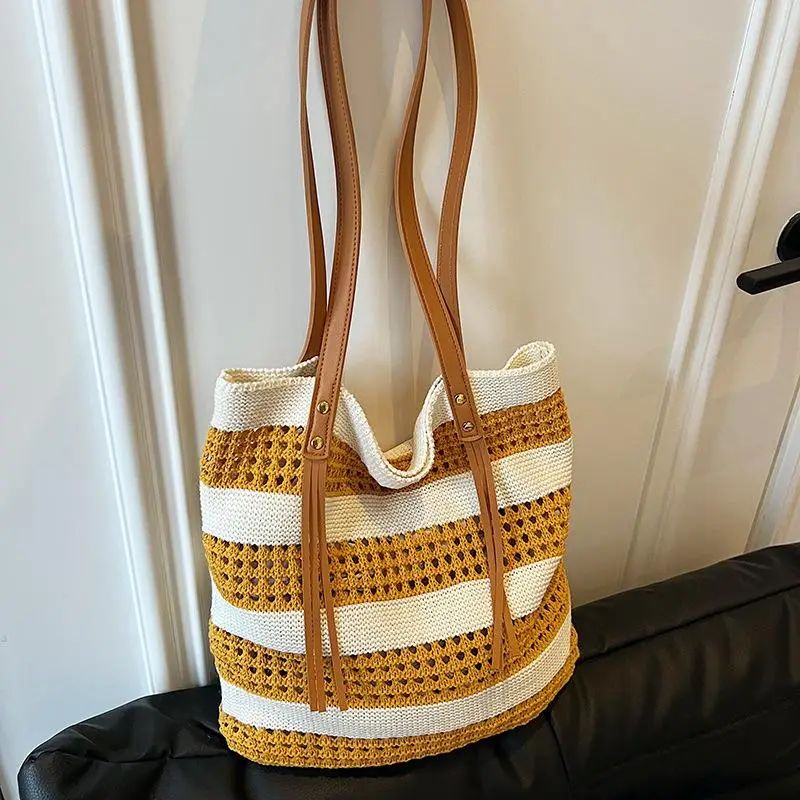 A woven straw tote bag with horizontal mustard yellow and cream stripes, featuring tan faux-leather shoulder straps, resting on a black leather sofa in front of a white paneled door.