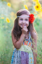 A young girl with long brown hair wearing a colorful patchwork dress and a red flower headband, posing in a sunflower field.