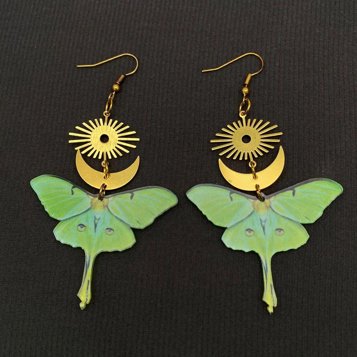 A pair of dangling earrings featuring light green luna moth wings with gold-tone sunburst and crescent moon charms above, displayed on a dark textured background.