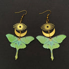 A pair of dangling earrings featuring light green luna moth wings with gold-tone sunburst and crescent moon charms above, displayed on a dark textured background.
