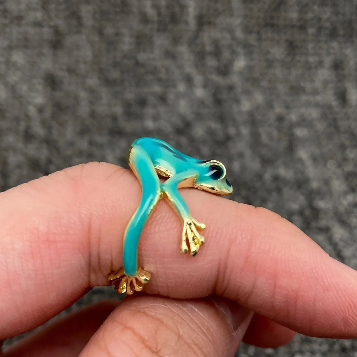 Turquoise and gold frog-shaped ring on a finger with a gray background