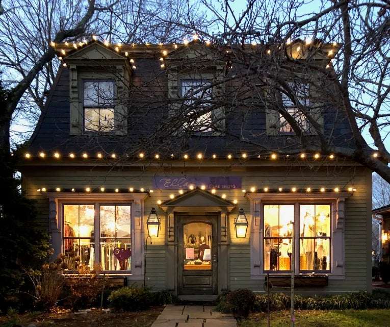 A charming Victorian-style house facade at dusk, decorated with warm string lights around the roofline and windows. A stone walkway leads up the front yard to the central door, flanked by grass and bare winter trees.