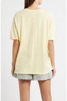 A woman shown from the back wearing a pale yellow oversized short-sleeve top with a vertical seam detail and striped shorts against a light gray studio background.