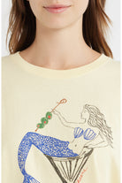 A close-up of a model wearing a cream-colored t-shirt with a graphic of a mermaid in a martini glass holding a skewer of green olives and red peppers. The brand name 'Sourpuss' is printed in red script near the bottom right of the graphic.