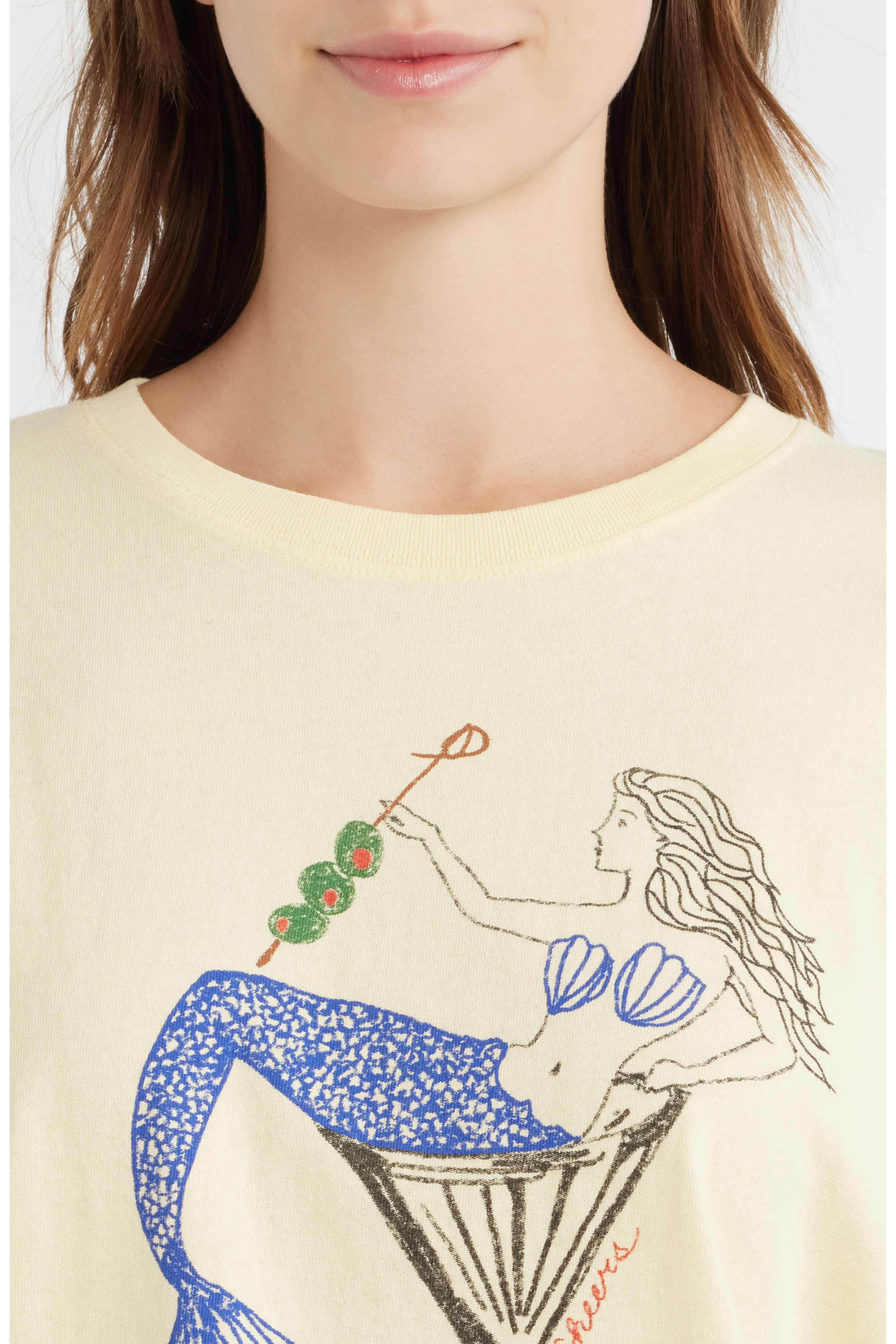 A close-up of a model wearing a cream-colored t-shirt with a graphic of a mermaid in a martini glass holding a skewer of green olives and red peppers. The brand name 'Sourpuss' is printed in red script near the bottom right of the graphic.