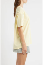 A woman shown in side profile wearing an oversized pale yellow short-sleeve T-shirt and striped shorts against a plain light gray studio background.
