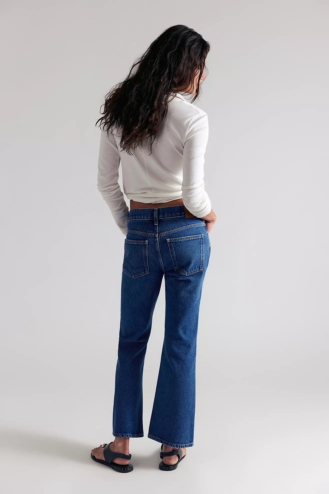 A woman shown from behind wearing blue flare jeans, a white long-sleeve top, and black sandals against a light gray studio background.