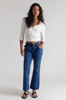 A woman modeling a white cropped long-sleeve henley top and blue high-waisted flare jeans with a black belt, paired with black sandals, against a light gray studio background.