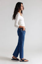 A woman in a white long-sleeve top and blue jeans stands in profile against a light gray studio background, wearing black sandals and a black belt.