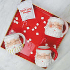 White and red ceramic Santa mugs on a red tray with mini marshmallows, candy canes, and hot cocoa